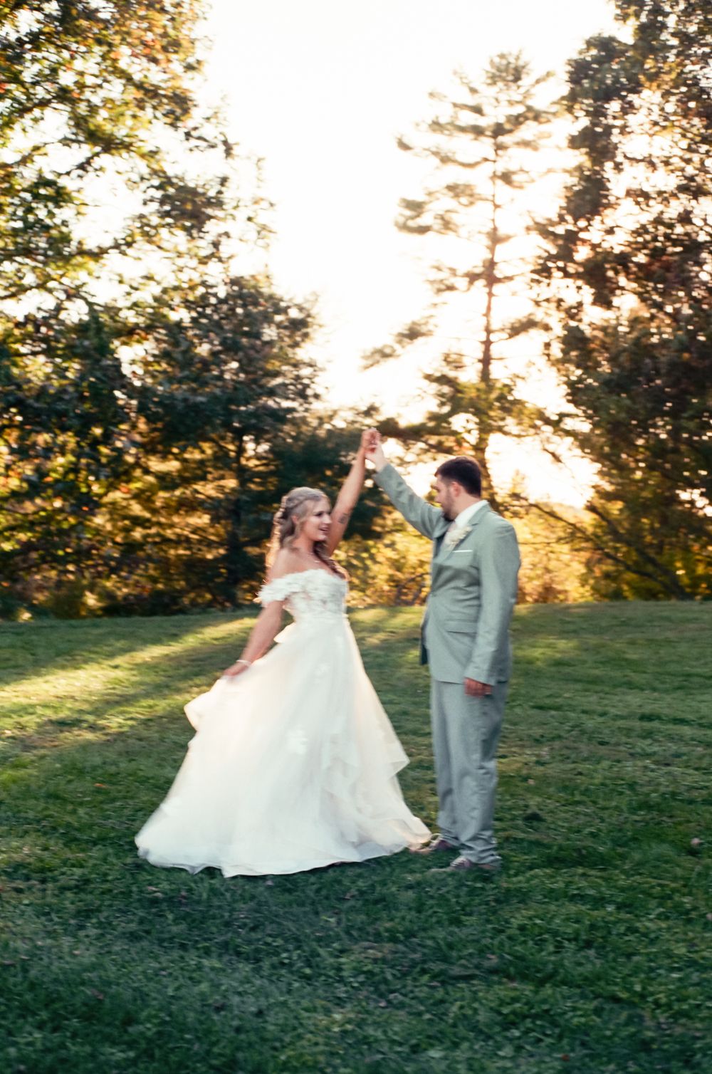 Wedding couple dances playfully in outdoor setting with sunset through trees.