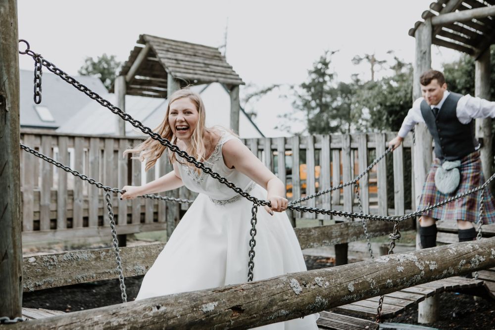 A bride in a white dress playfully poses on a wooden bridge with chains during a wedding photo session.