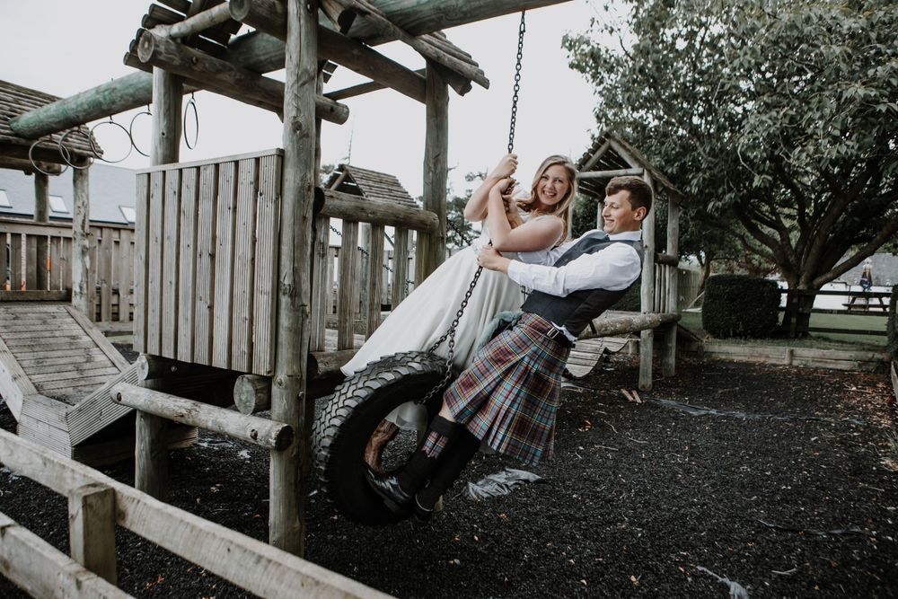 A couple share a playful moment on a wooden swing set in a rustic outdoor setting.