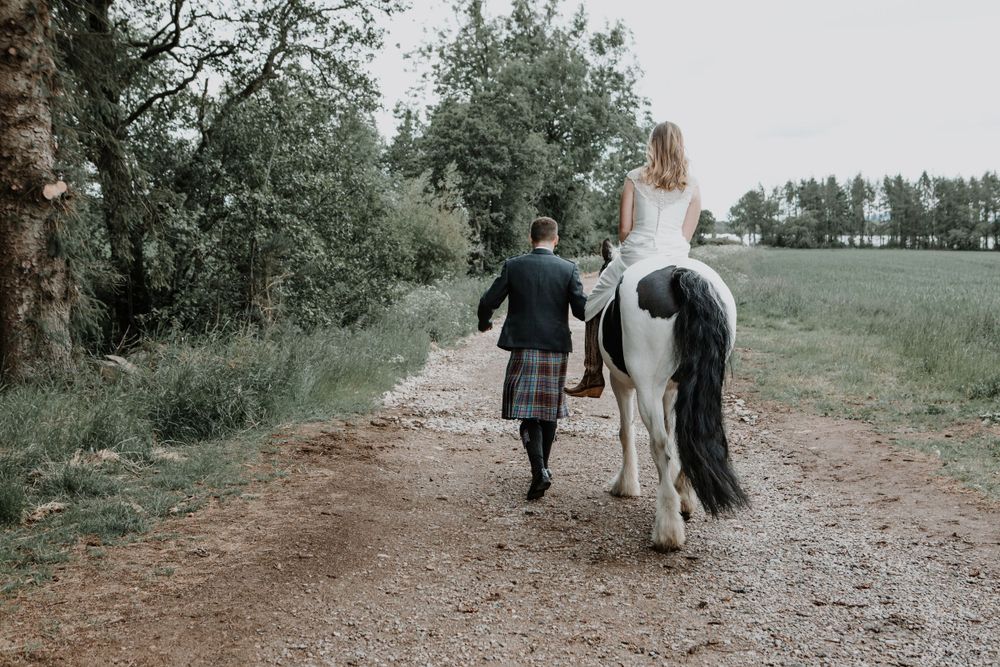 A black and white horse carries a rider down a country path while someone walks alongside.