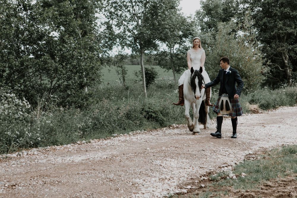 A couple in vintage wedding attire walk together with a white horse on a country path.