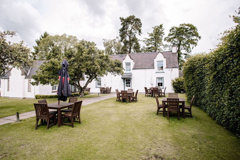 Garden setup with wooden picnic tables and chairs on a lawn surrounded by white buildings and trees.