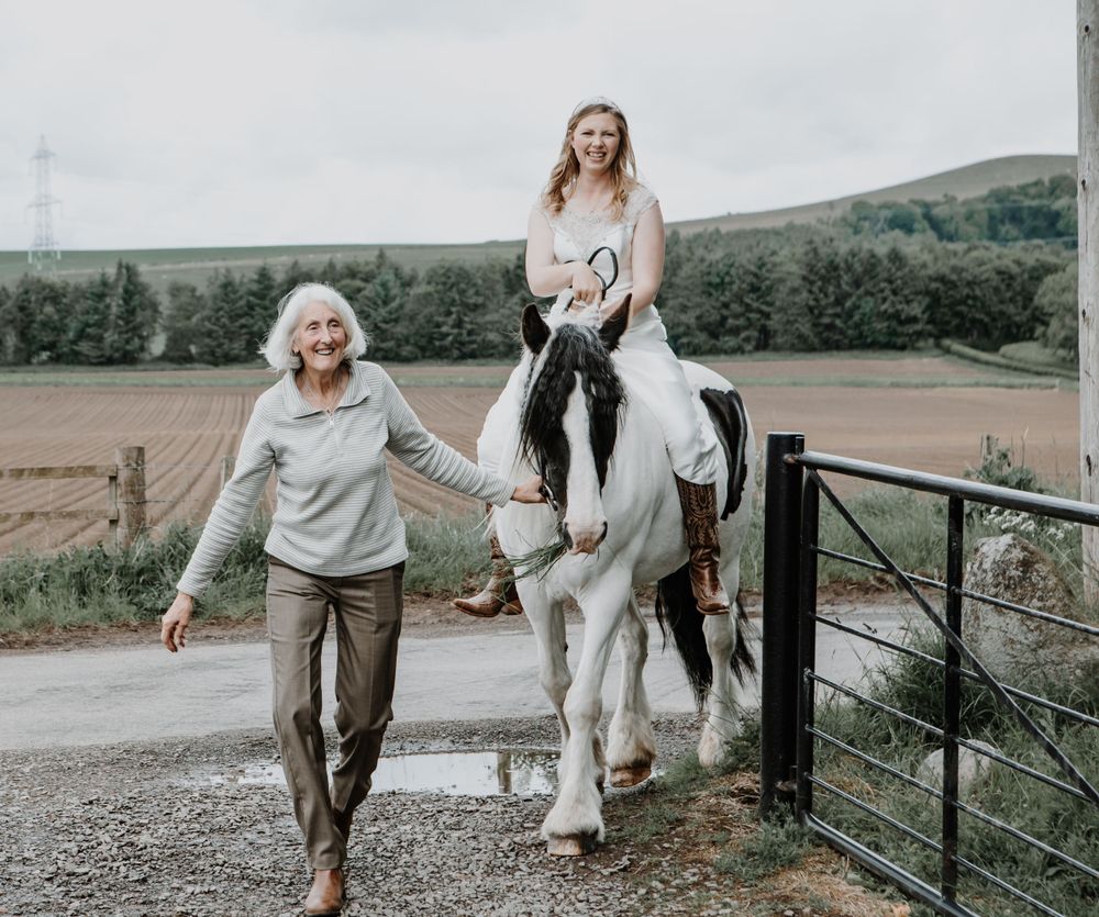 A white horse carries a rider along a country path while someone walks alongside on a cloudy day.