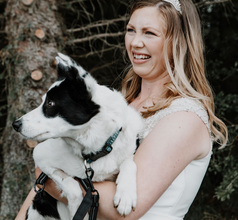 A person in a white top smiles while holding a happy black and white dog outdoors.