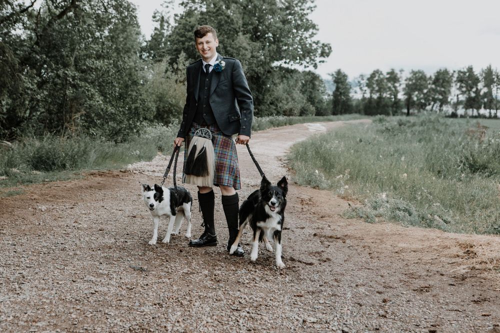 A person in formal Scottish attire stands on a dirt path with two black and white dogs.