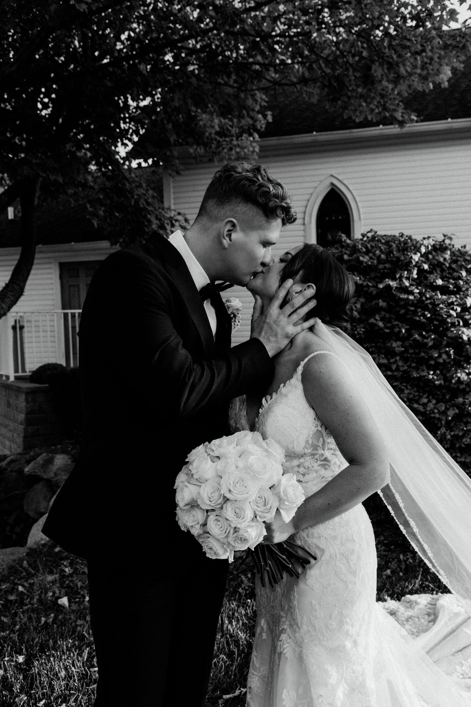 A wedding couple shares a kiss outdoors near a white chapel with the bride holding a bouquet of white roses.