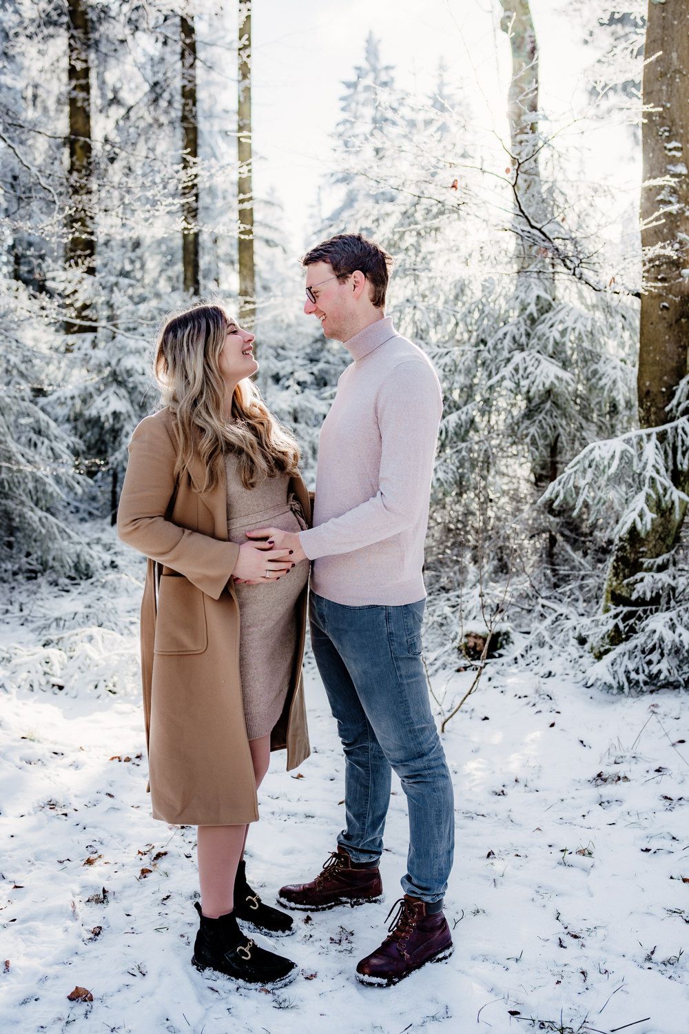 A couple embraces in a snowy winter forest setting during a maternity photoshoot.