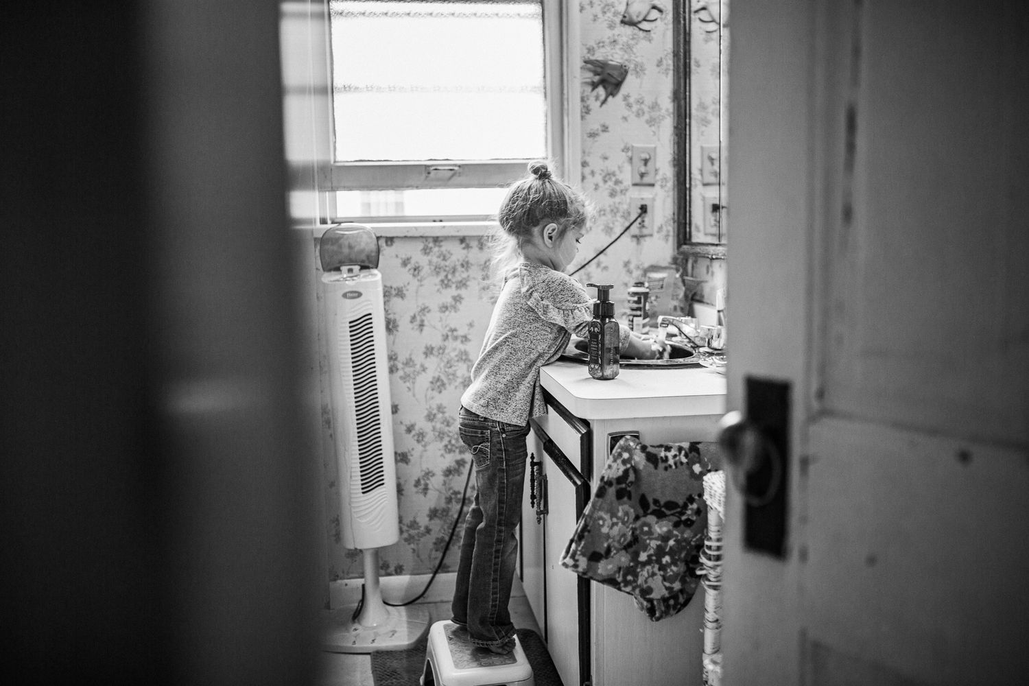 A child stands on a stool at a kitchen counter in a black and white photograph taken through a doorway.