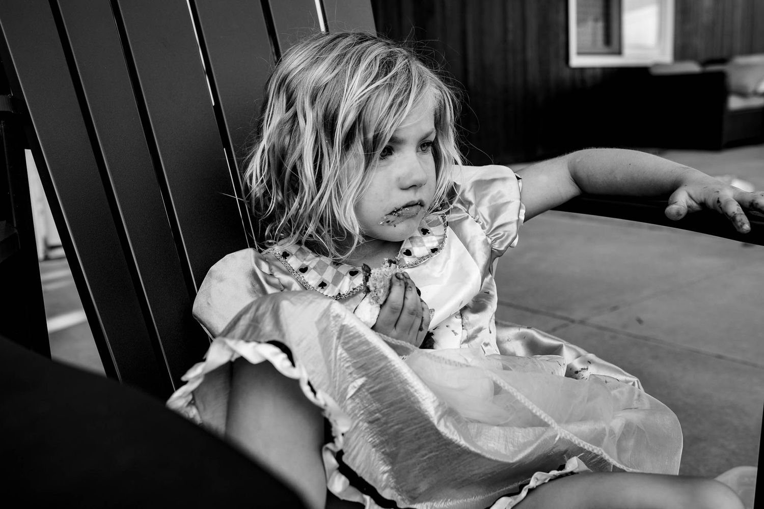 Black and white photo of a young child in a ruffled dress lounging on a vintage chair at a dining table.