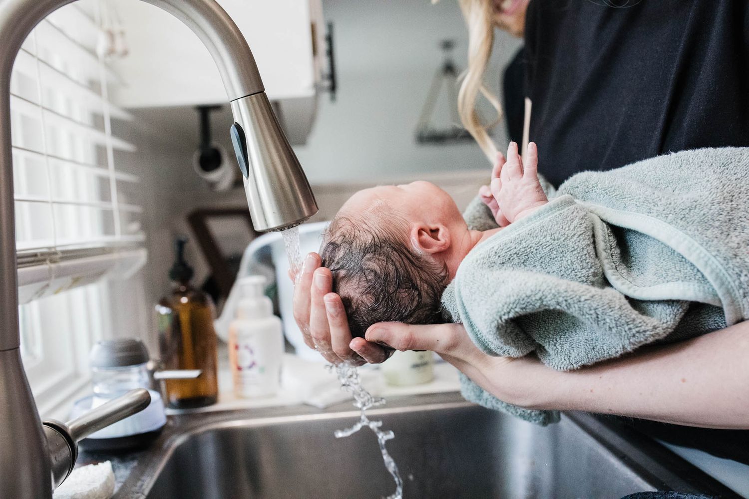 Baby being bathed in kitchen sink while cradled in someone's arms with blue towel.