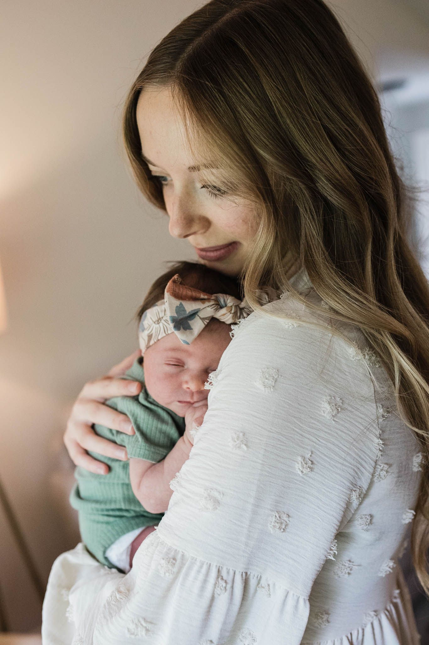 A tender moment shows a woman in a white dress holding a sleeping newborn baby wrapped in mint green.