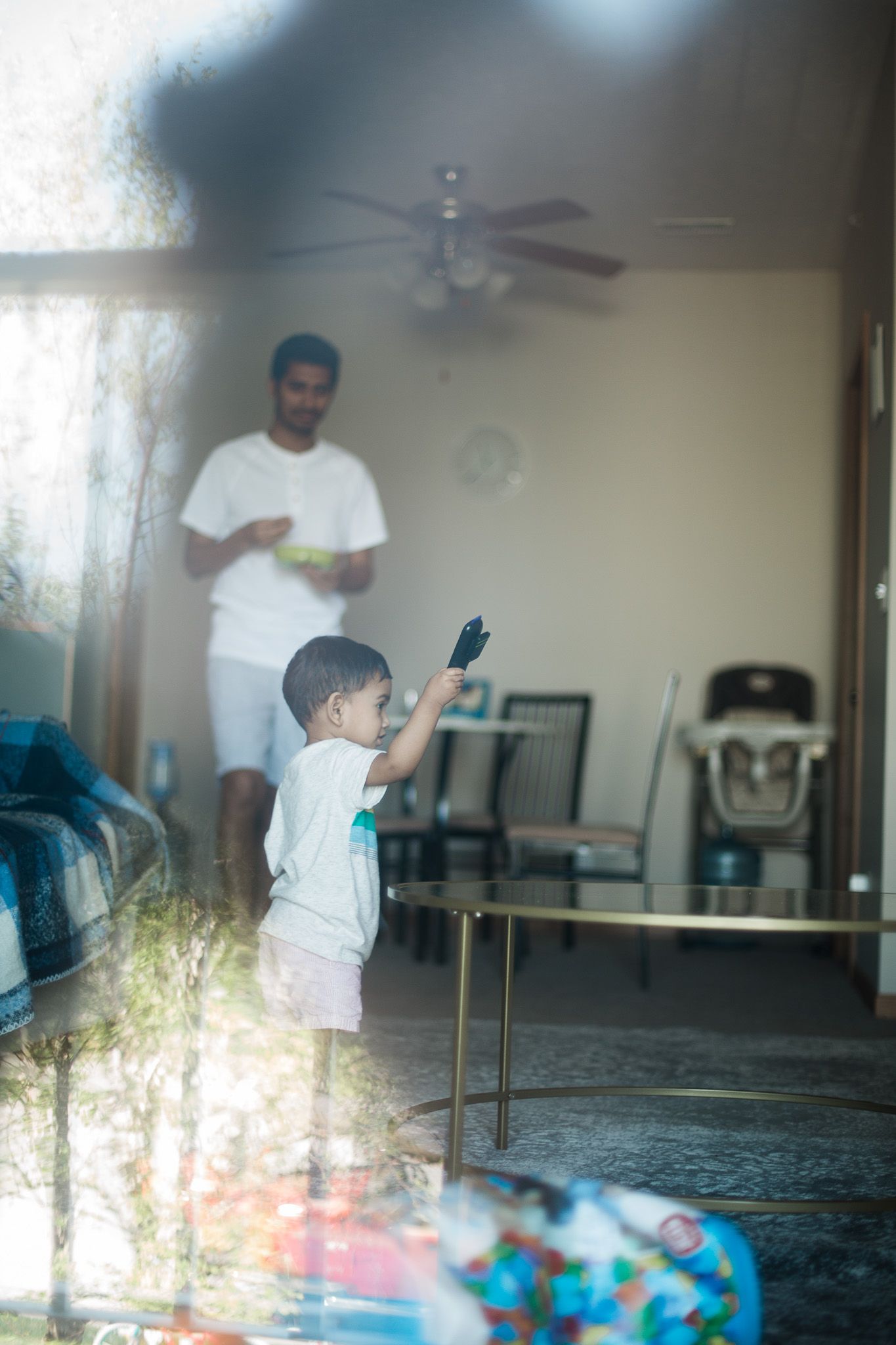 Through a window reflection, a young child stands with raised arm in an indoor living space with a ceiling fan.