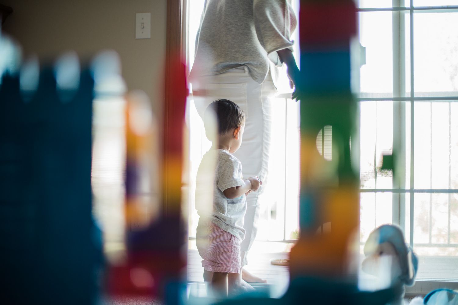 Through colorful building blocks in the foreground, a figure in white reaches for a window curtain in a bright room.