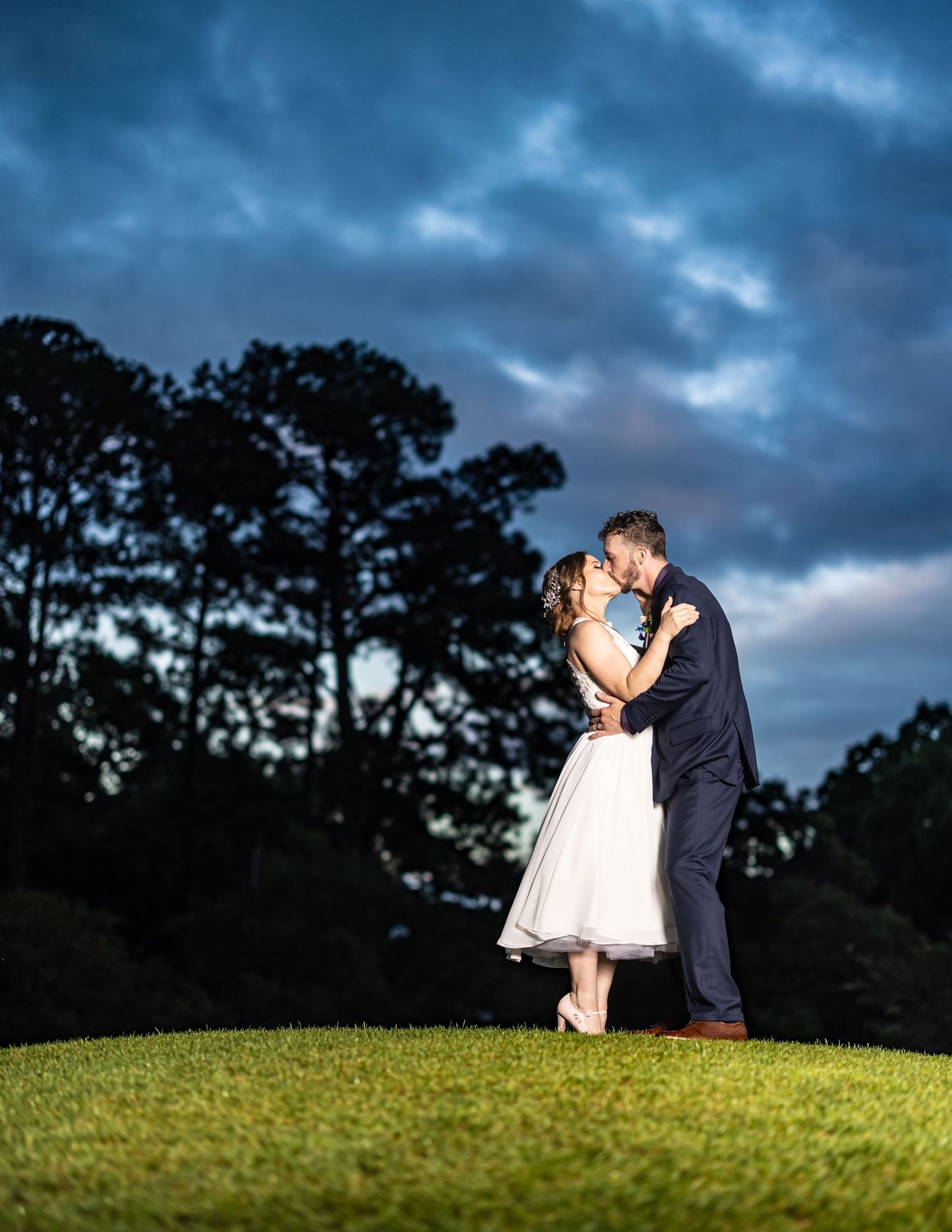 A romantic couple shares an intimate moment on a hilltop at dusk with dramatic stormy clouds in the background.