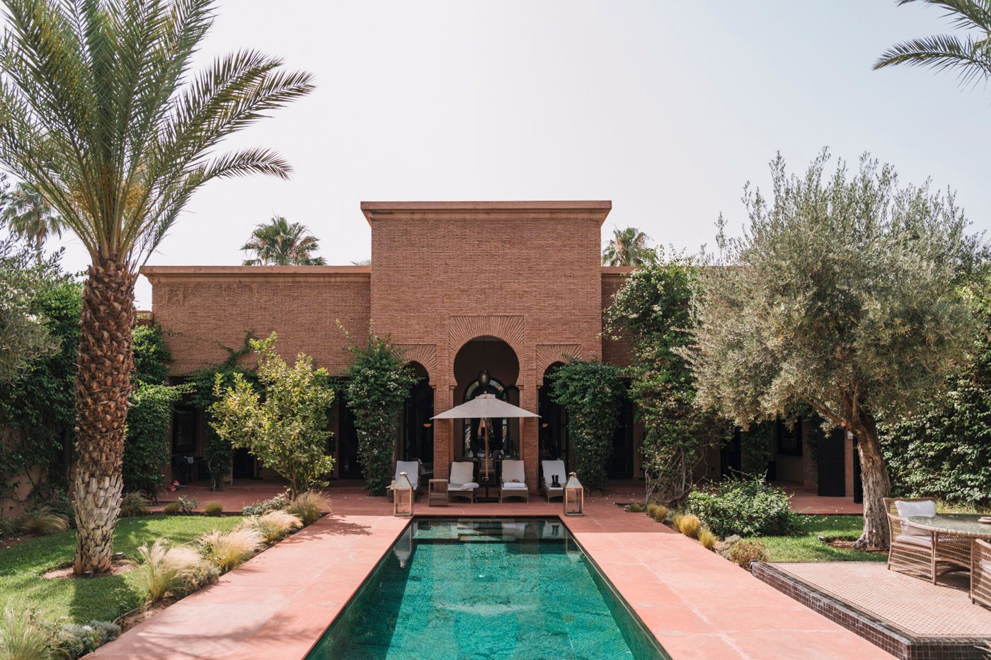 Luxurious Moroccan villa with terracotta walls, arched entrance, and rectangular swimming pool surrounded by palm trees.