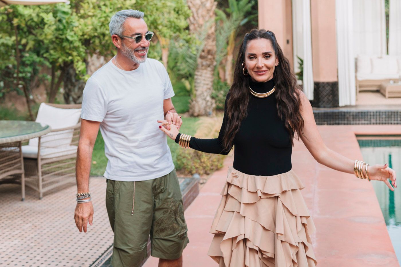 A pair walking outdoors on a patio, with one wearing a gray t-shirt and another in a flowy beige ruffled skirt.