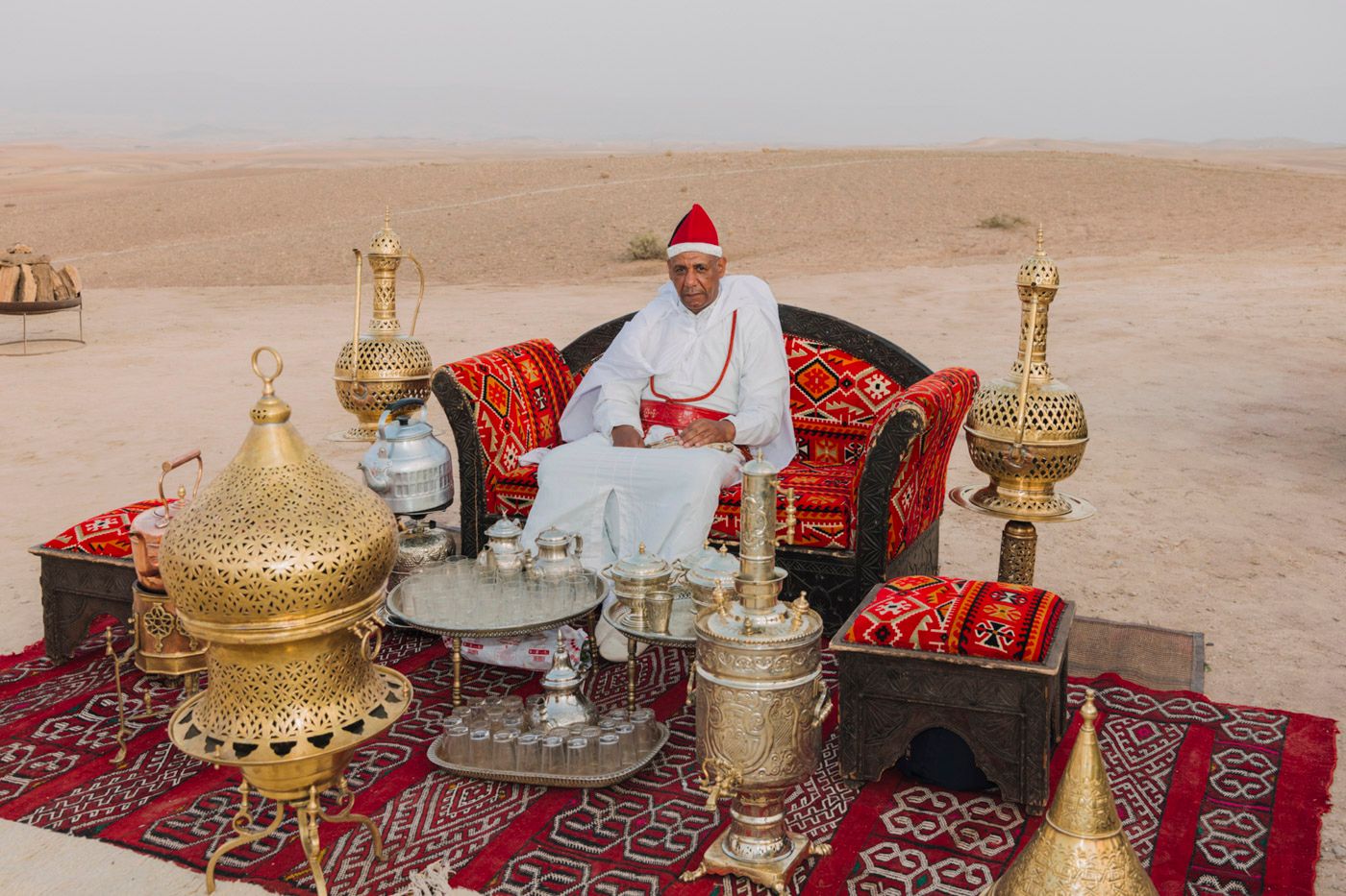 Traditional Moroccan tea service setup with brass teapots and ornate vessels displayed on vibrant red carpets in desert setting.