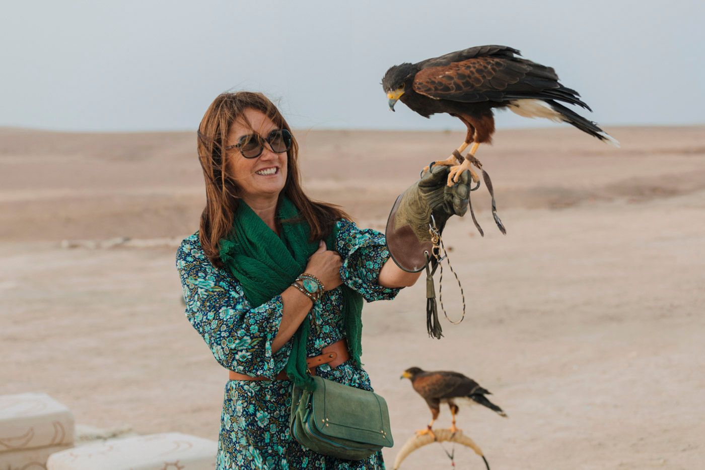 A falconer in a green patterned dress holds a hooded hawk in the desert as another bird perches nearby.