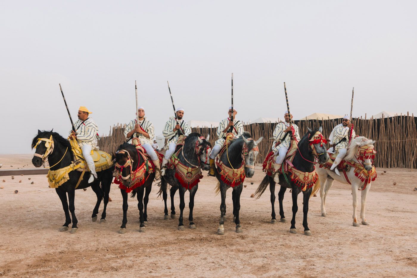 Row of mounted performers in traditional Moroccan attire on horseback in a desert setting.