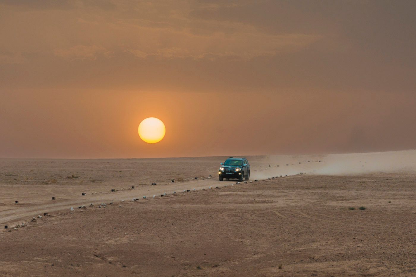 A turquoise 4x4 vehicle drives across a desert landscape during a golden sunset.