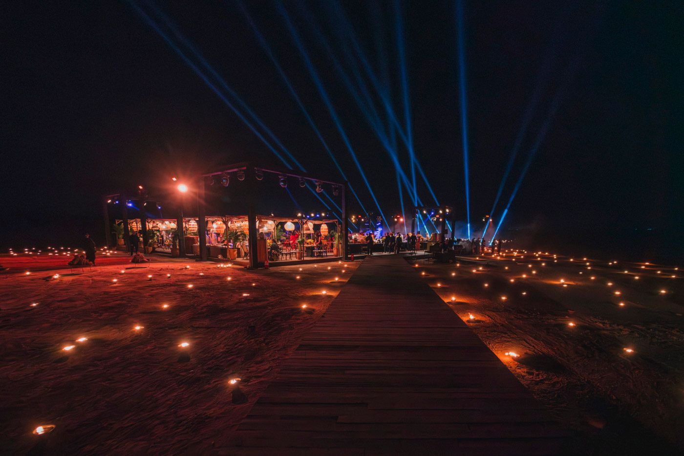 Illuminated wooden pier at night with glowing candles leading to a lit structure and blue searchlights in the sky.