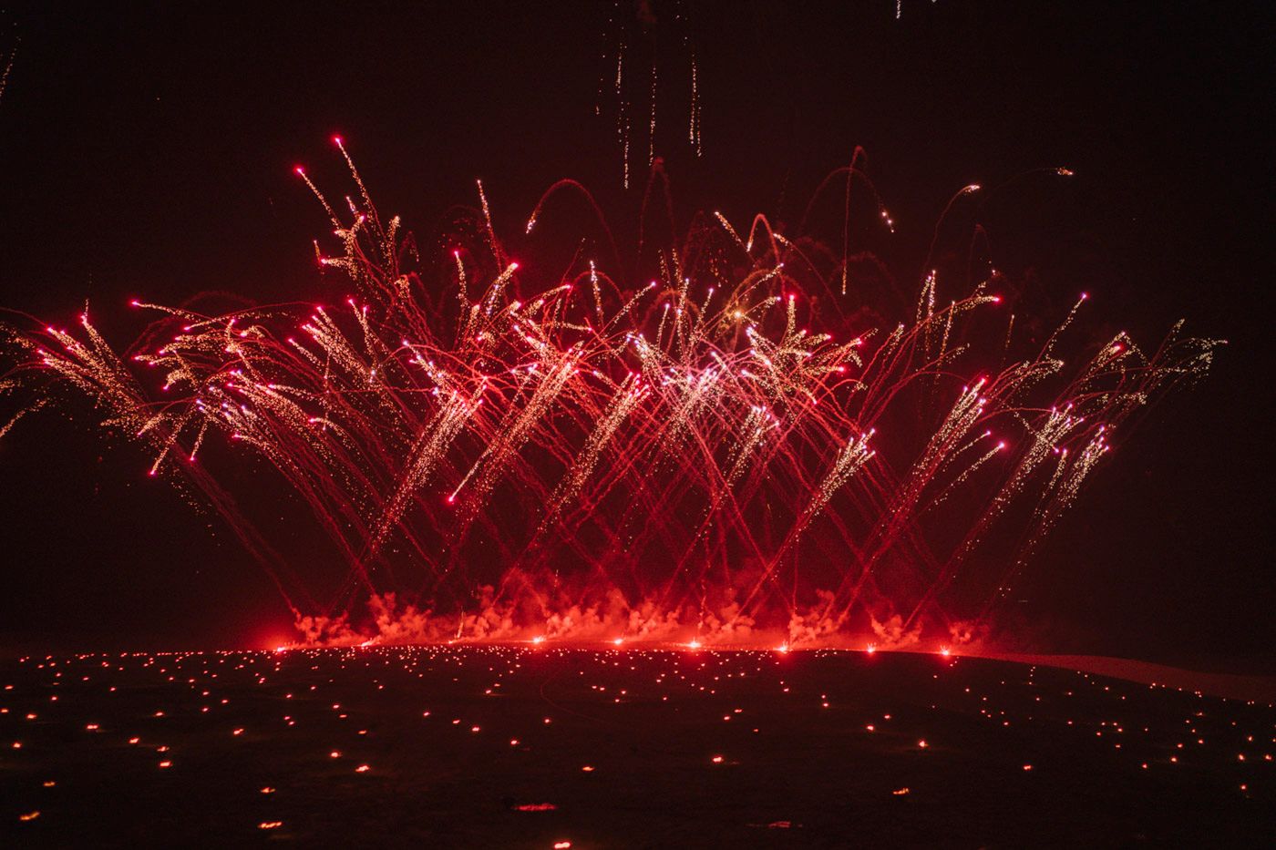 Bright red fireworks illuminate the night sky with dramatic bursts reflected on a dark surface below.