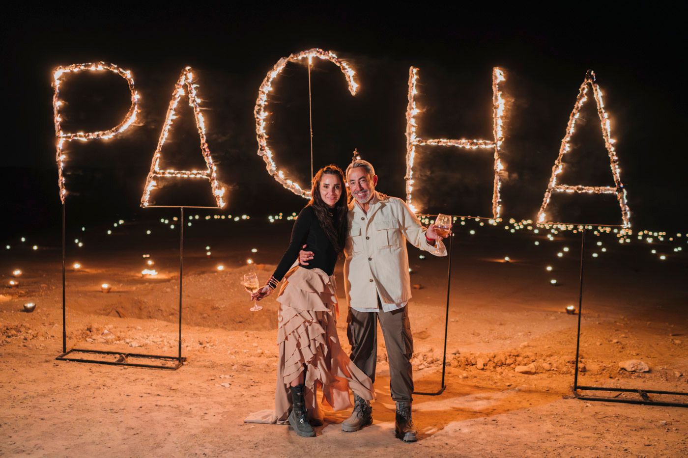 Two people standing in front of illuminated PACHA letters made of lights at night in a desert setting.