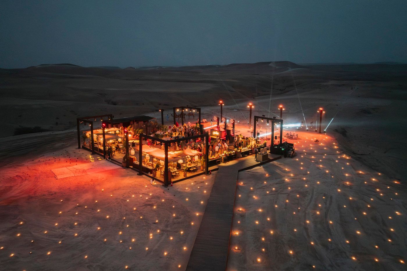 Glowing red lanterns illuminate a desert campsite at night with atmospheric lighting and cozy seating arrangements.