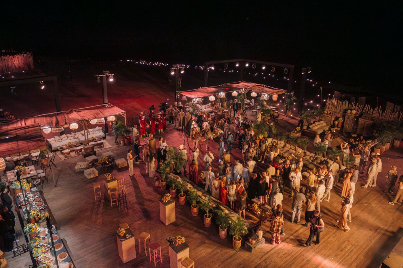 Aerial view of a crowded outdoor nighttime event with illuminated bars and gathering spaces on a deck.