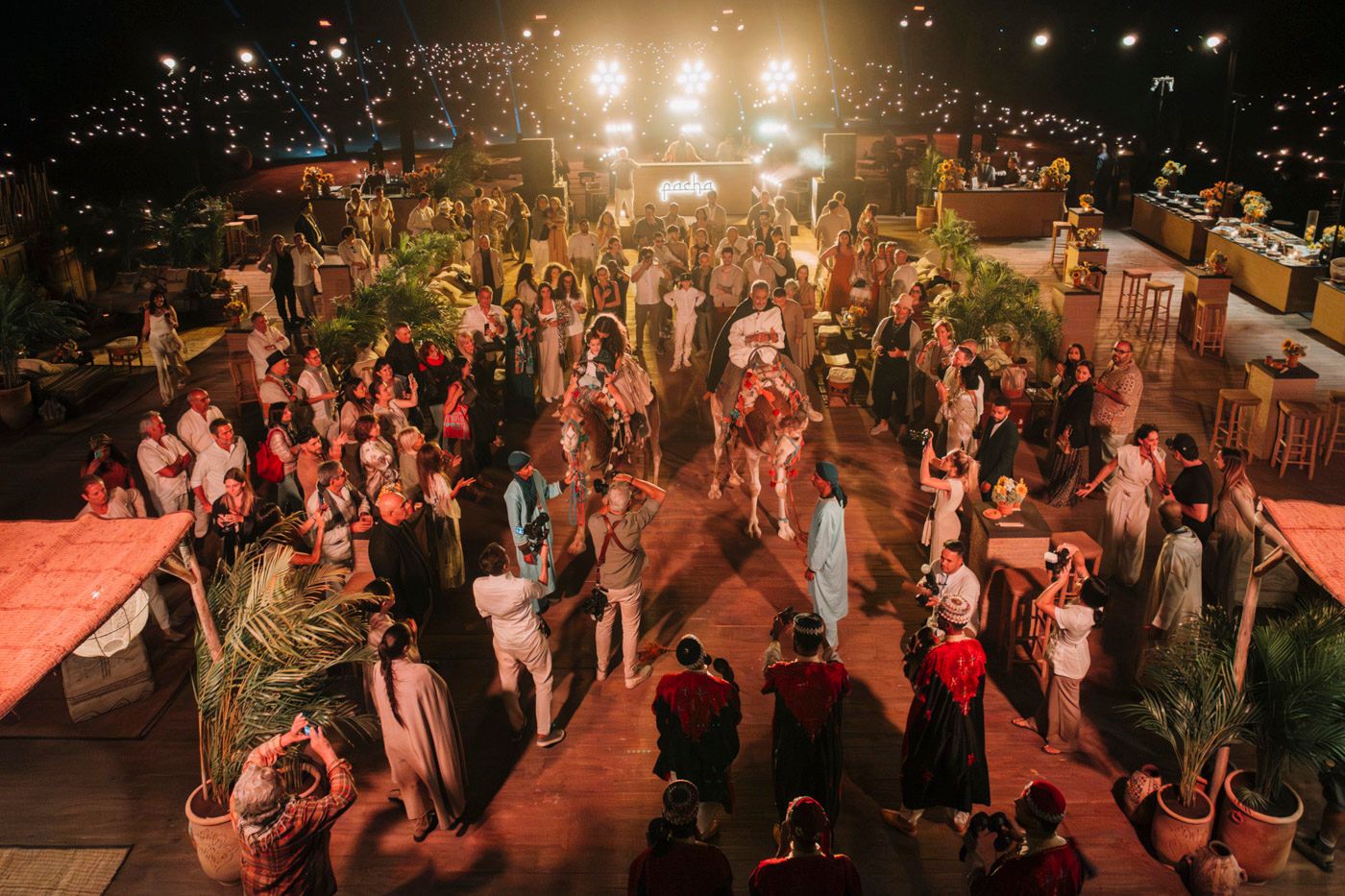 Large crowd gathered at an outdoor evening celebration with warm lighting and decorative elements.