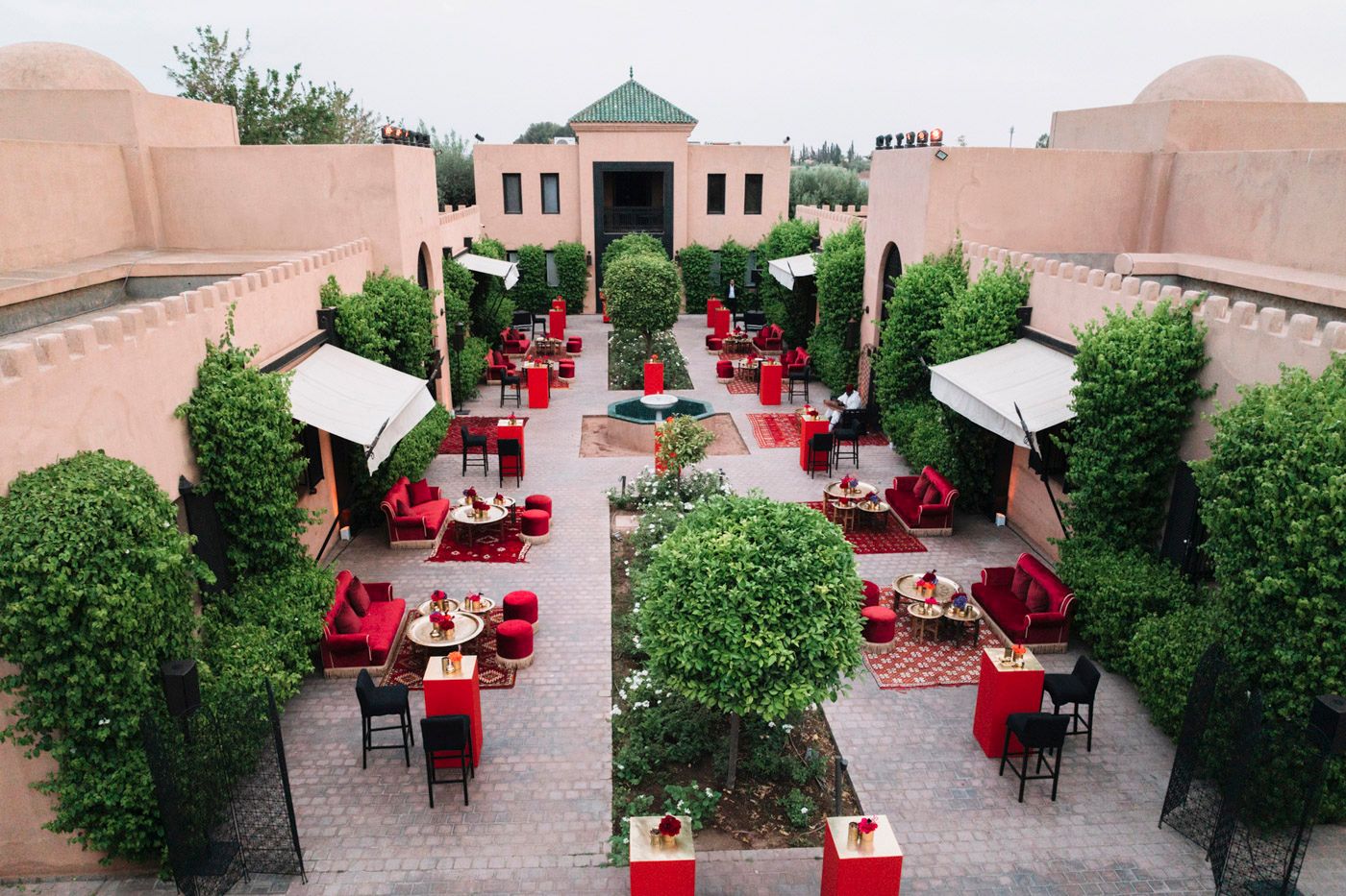 Aerial view of an open-air restaurant courtyard with red tables and chairs surrounded by pink walls and green landscaping.