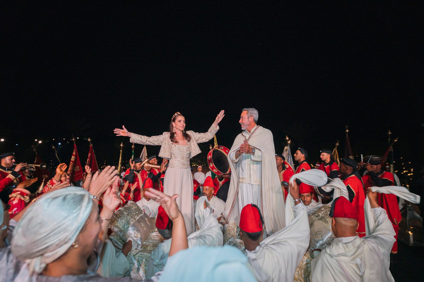 Bride and groom stand elevated among crowd of cheering guests in red and white attire at outdoor nighttime celebration.