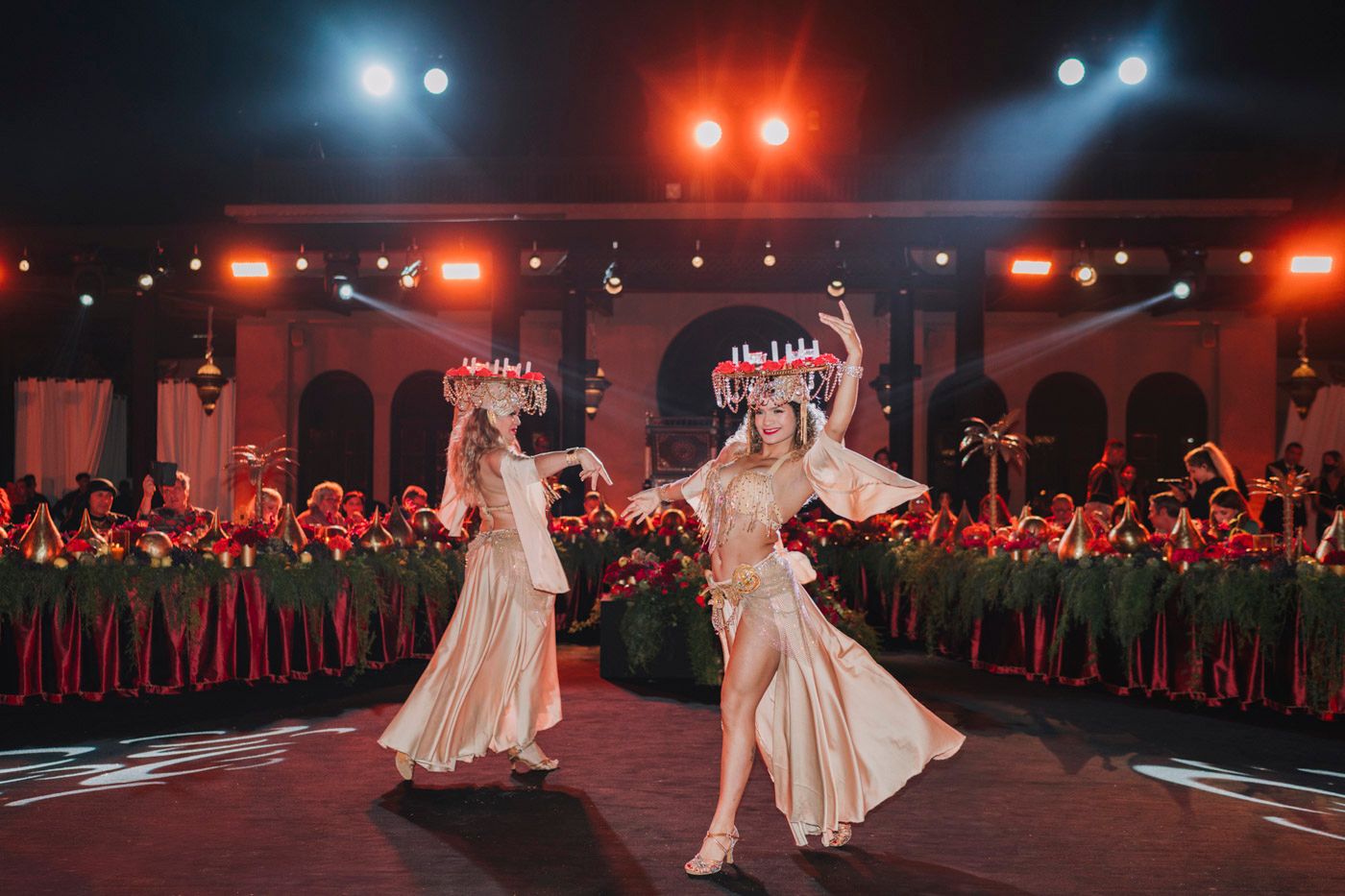 Two dancers in flowing white dresses perform on a circular stage surrounded by red floral arrangements under dramatic lighting.