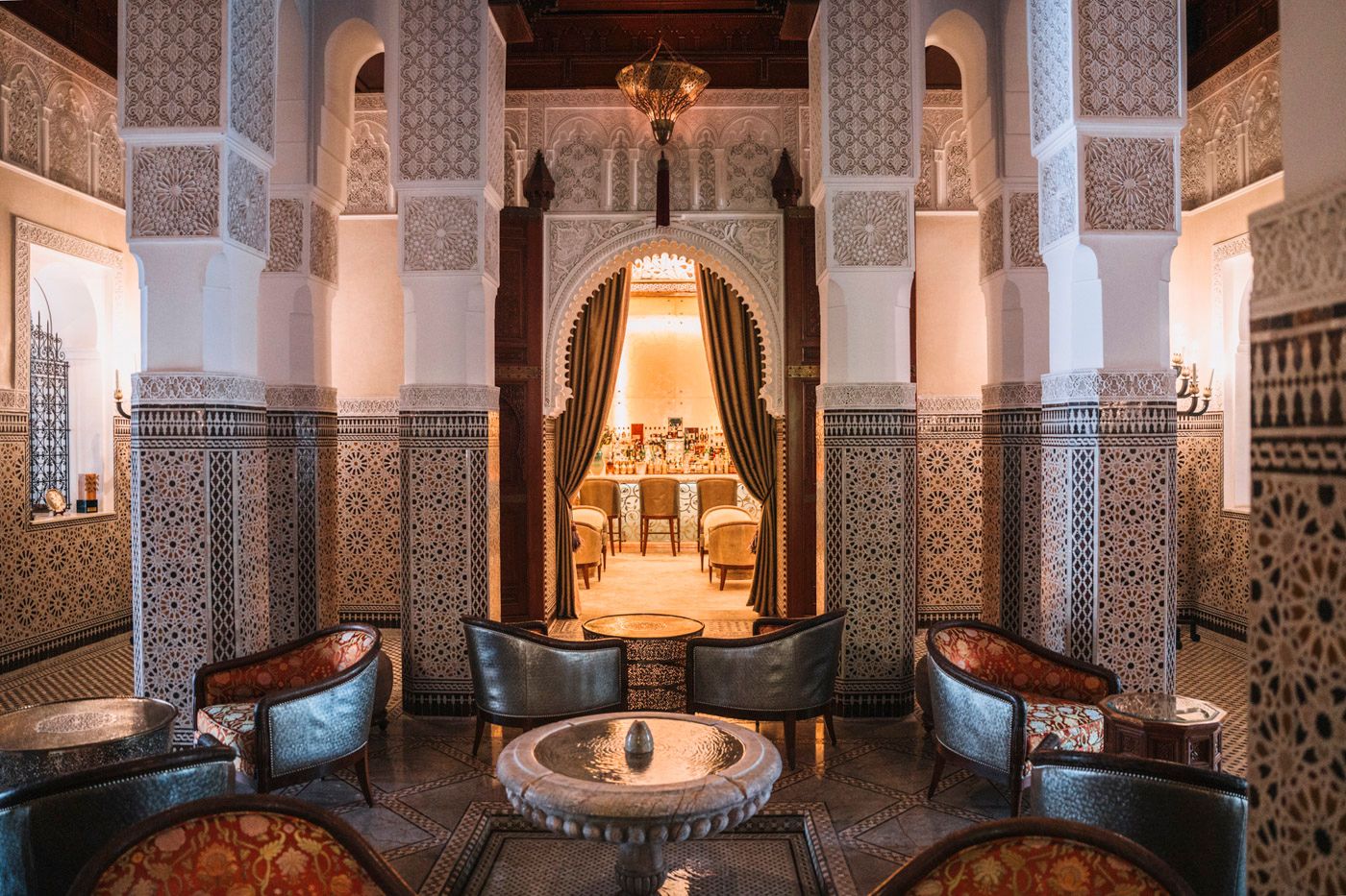 Luxurious Moroccan lounge with ornate archways, blue velvet chairs, and intricate tile work in a warm, lit riad interior.