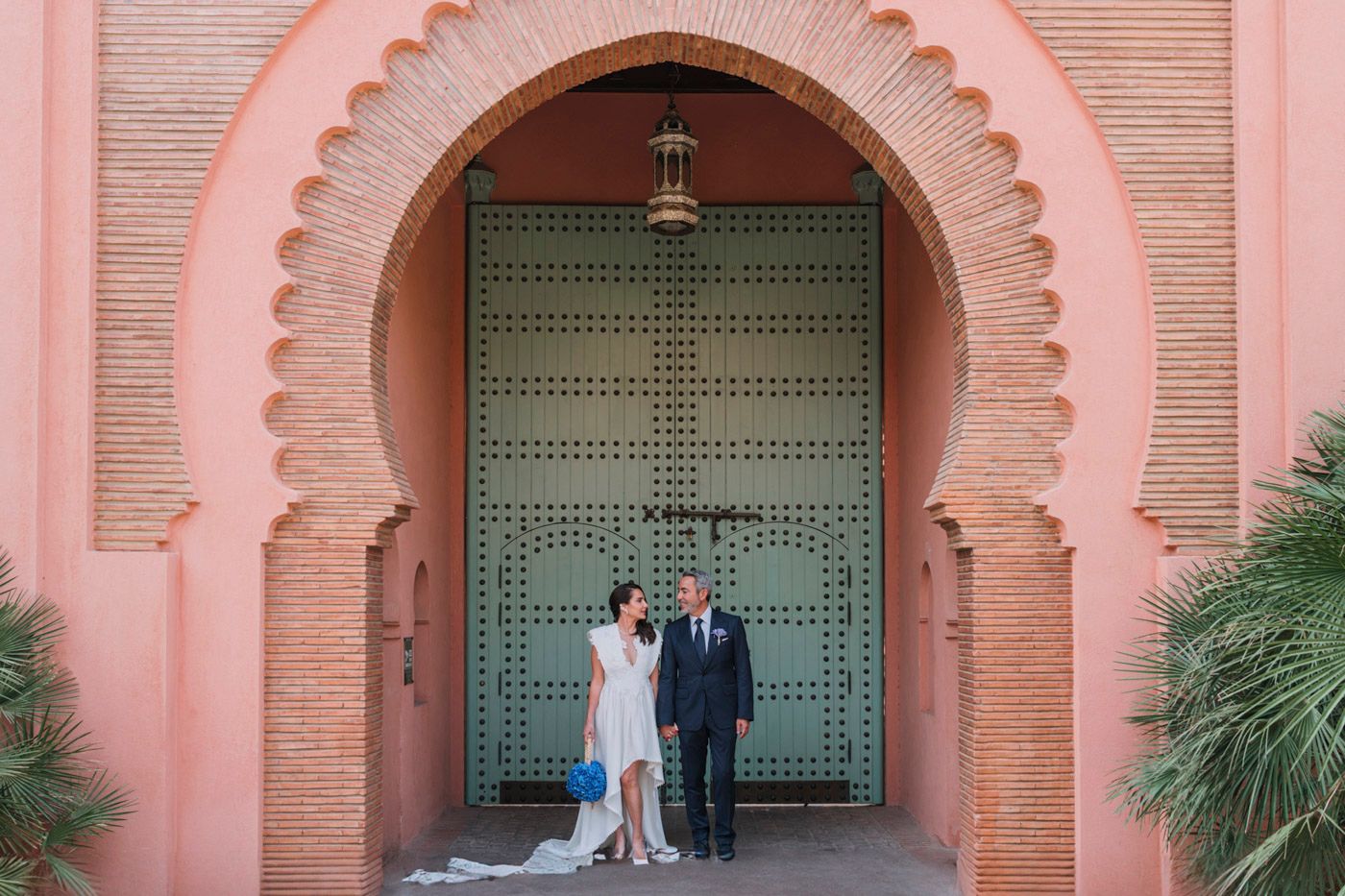 Two people stand in front of an elegant pink Moroccan-style archway with turquoise doors at a luxury resort.