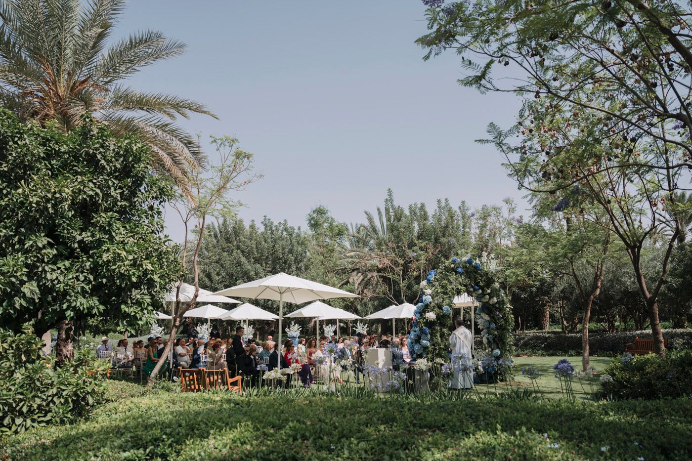 Outdoor garden party setup with white umbrellas and guests gathered among lush palm trees and greenery.