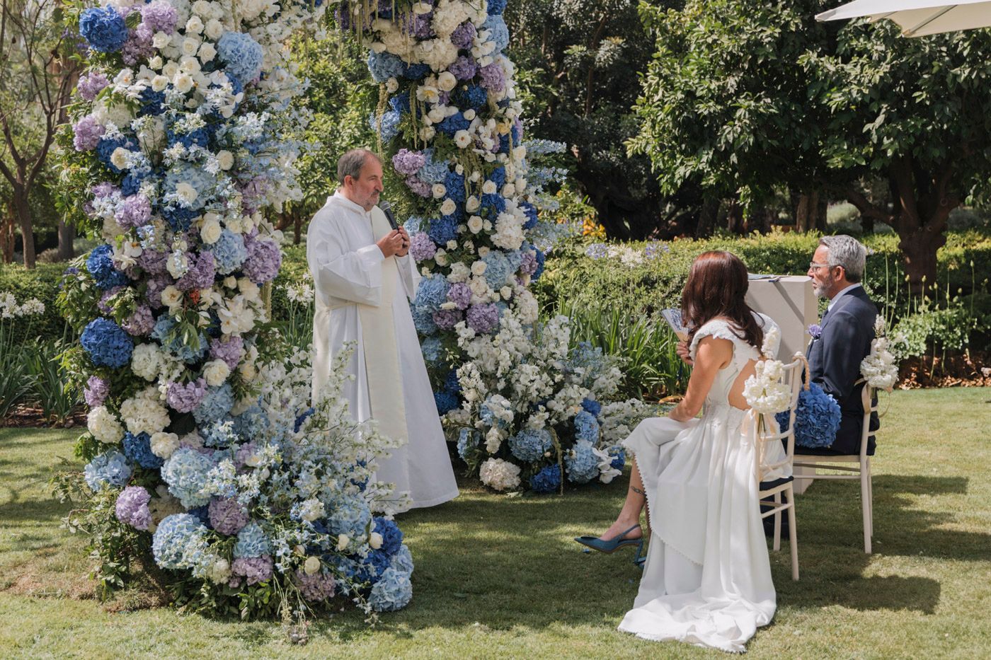 Outdoor garden wedding ceremony with blue and white floral arch and celebrant standing under flowering decorations.