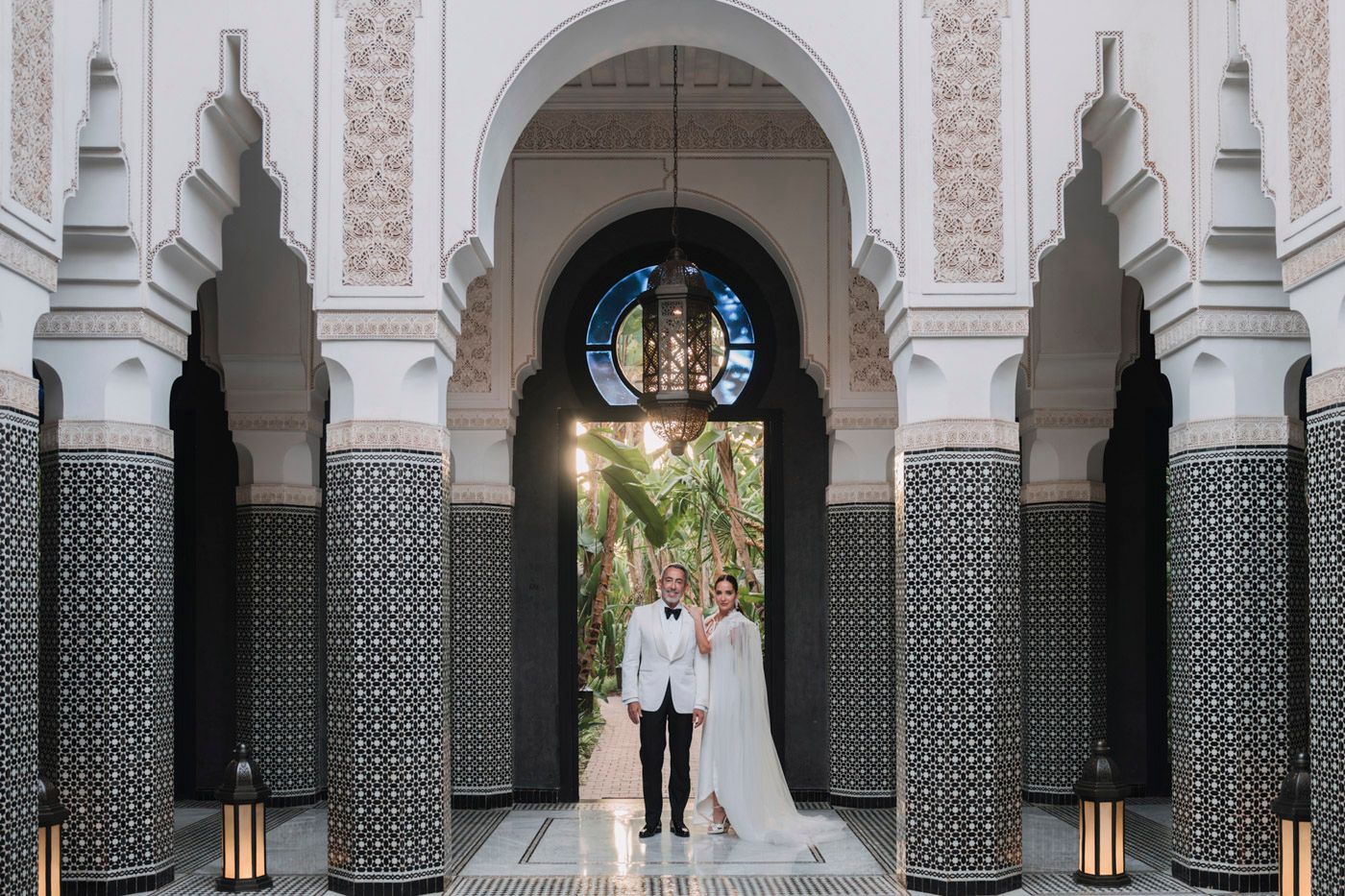 Couple stands beneath ornate Moroccan archway with geometric tiles and white columns at luxury resort.