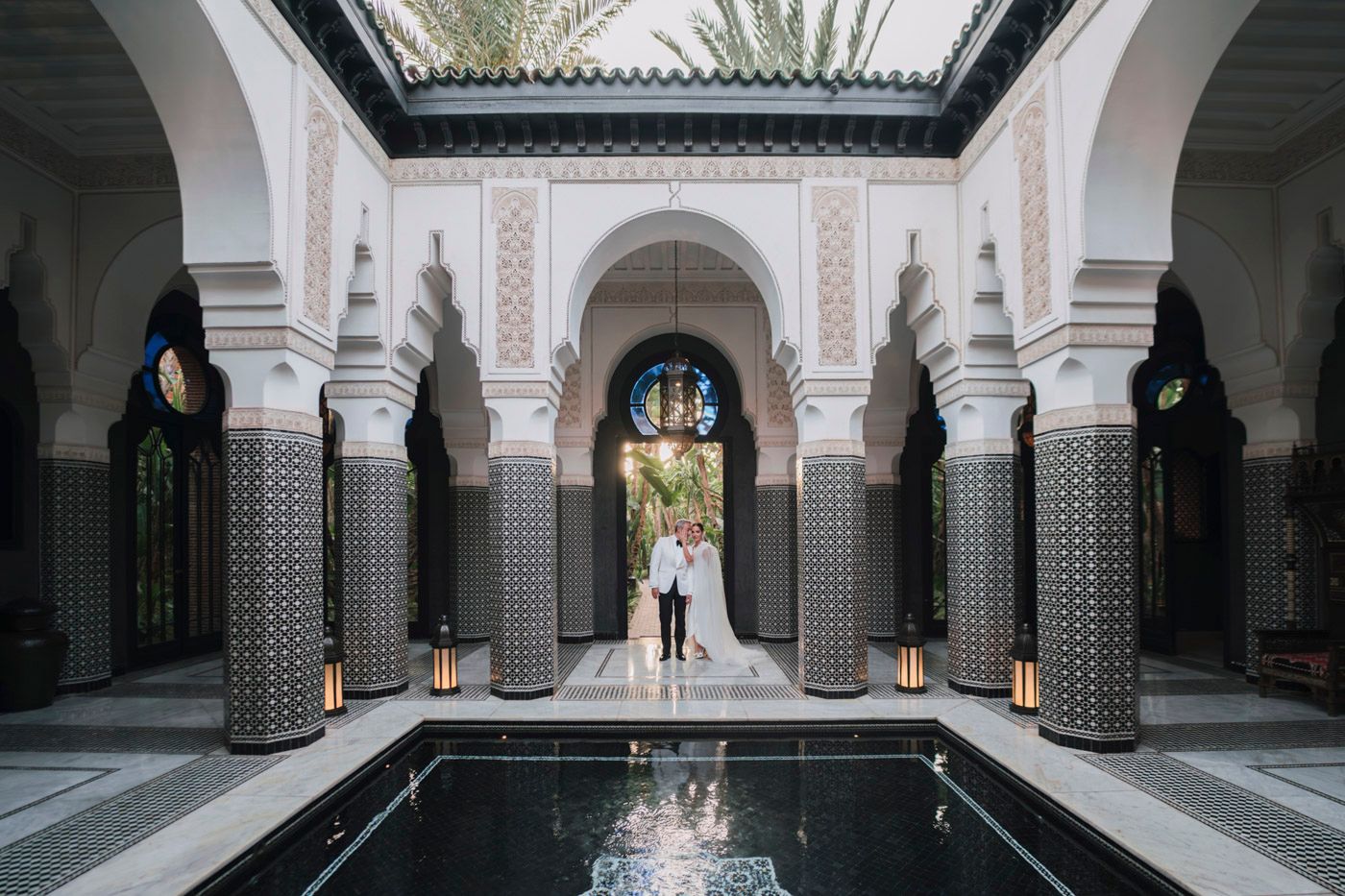 Elegant Moroccan riad interior with ornate columns, reflecting pool and traditional tilework surrounding a covered courtyard.