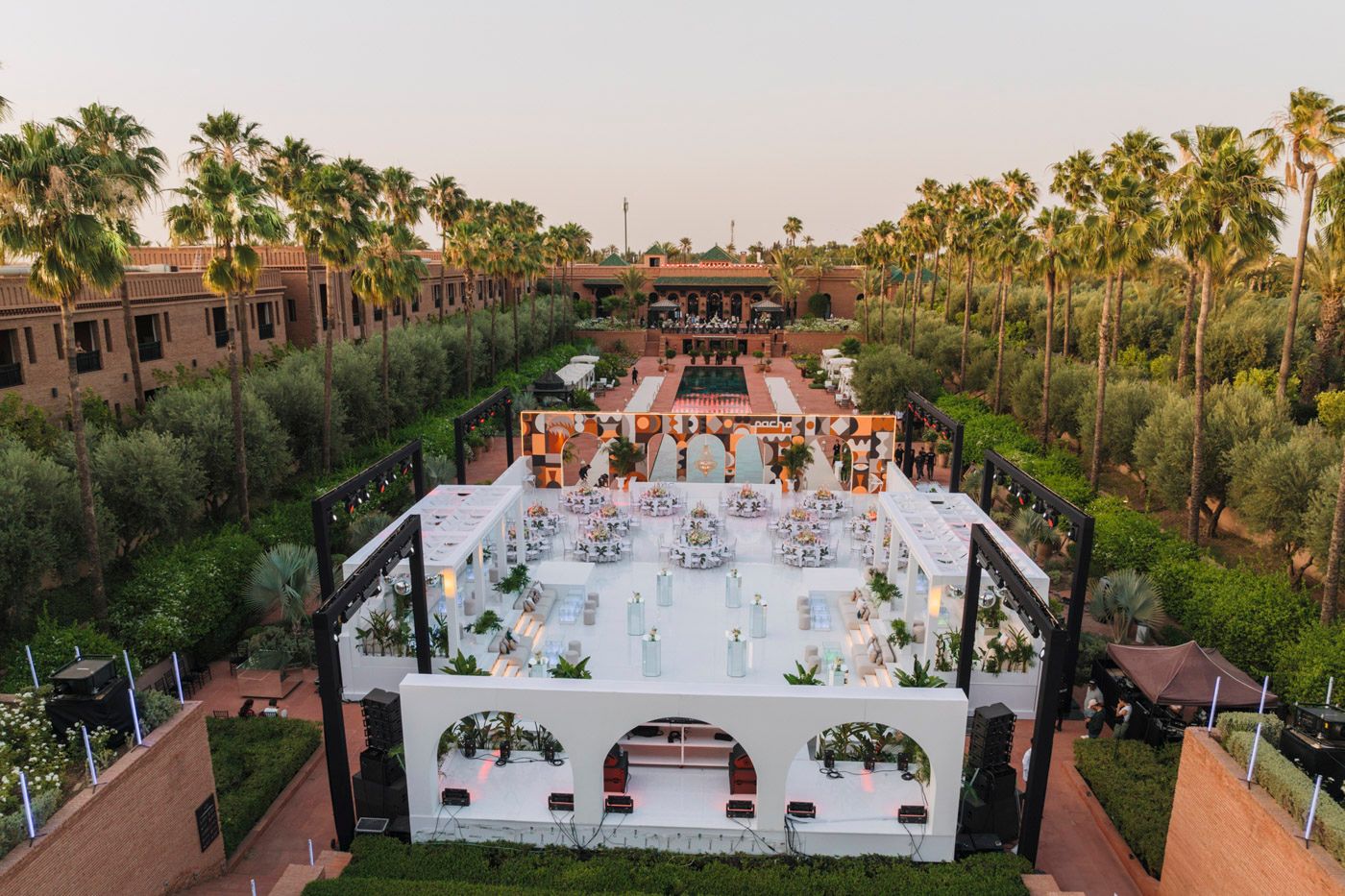 Aerial view of an elegant outdoor wedding reception setup with white dance floor surrounded by palm trees at dusk.