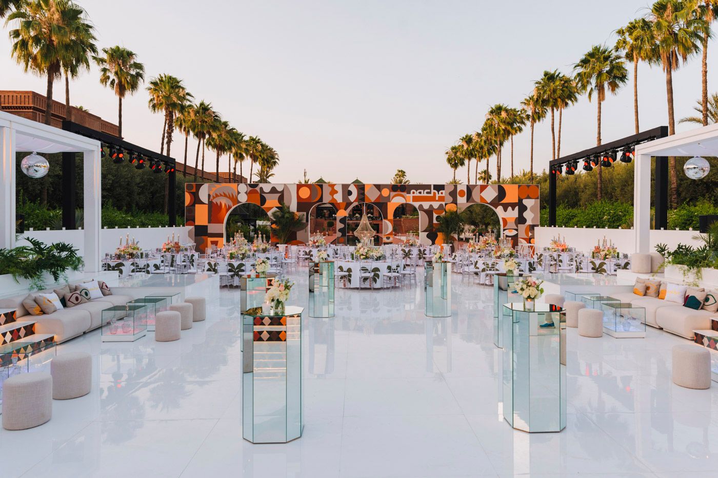 Elegant outdoor wedding reception setup with palm trees and white decor around a reflective pool at sunset.