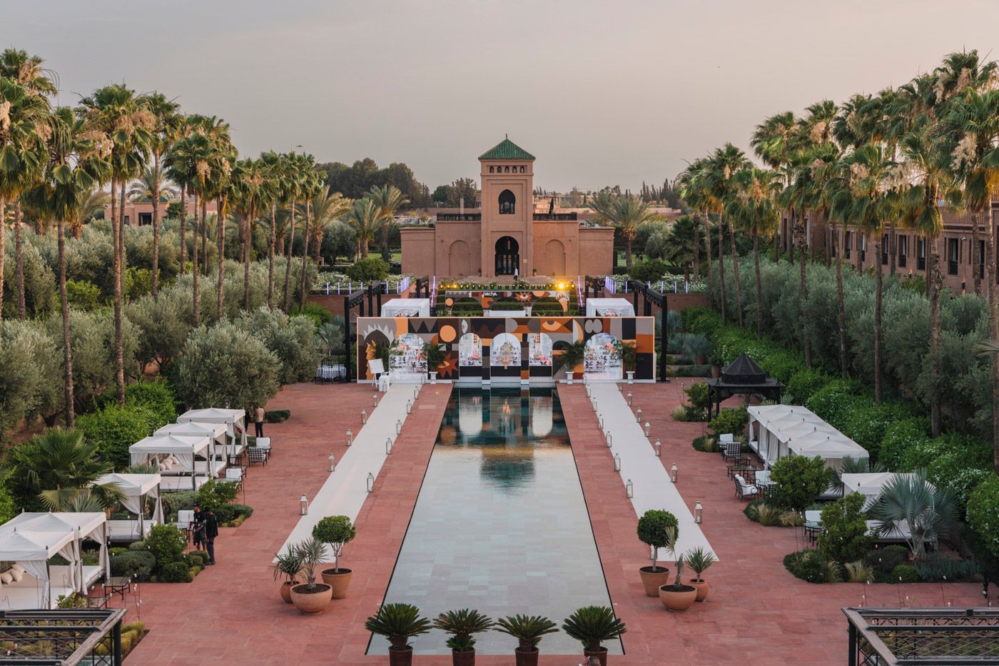 A luxurious Mediterranean resort courtyard with reflecting pool, lined with palm trees and outdoor seating at sunset.