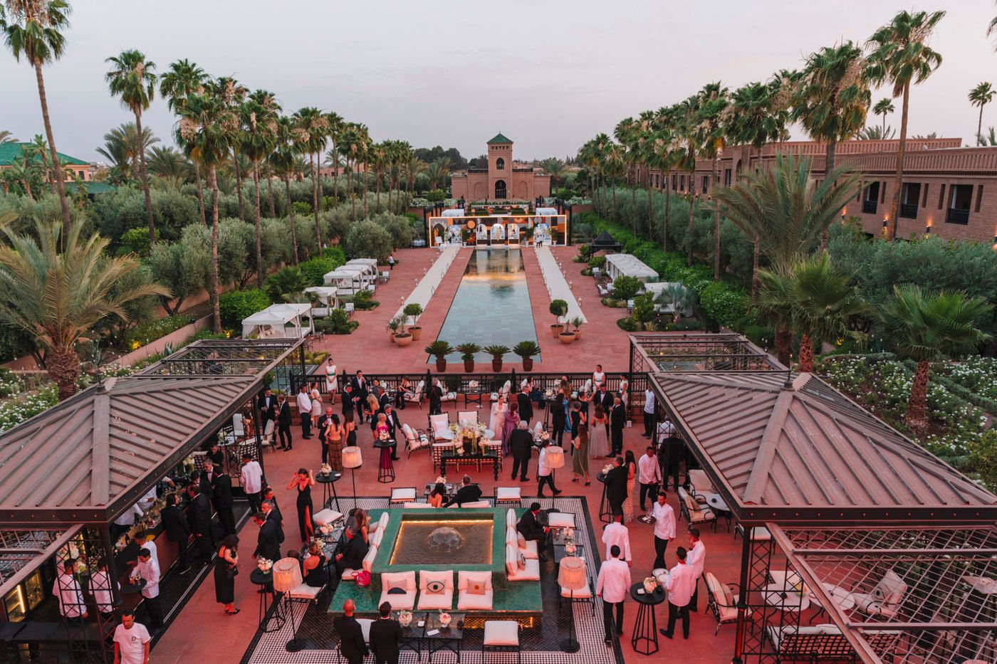 Aerial view of a luxury resort courtyard with reflecting pool, palm trees and crowd gathered for evening event.
