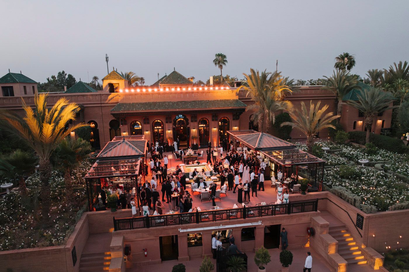 Aerial view of an outdoor evening wedding reception at a Mediterranean venue with palm trees and string lights.