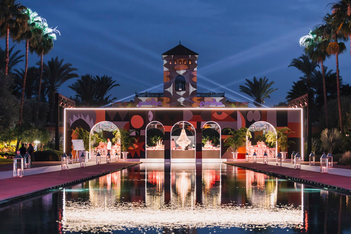 Illuminated oriental-style pavilion with pink lights reflects in a symmetrical pool at twilight, surrounded by palm trees.