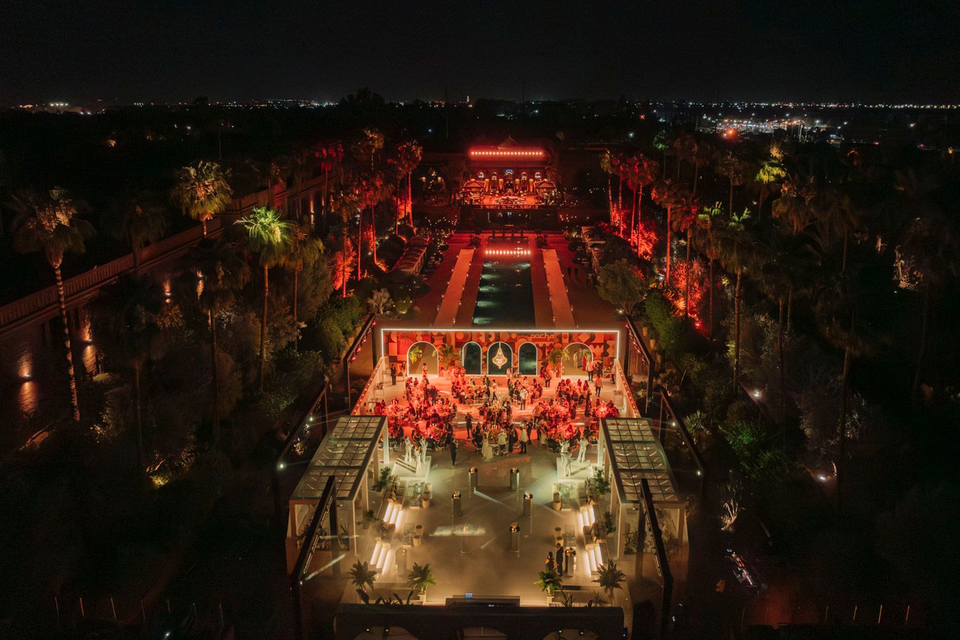 Aerial view of an elegant outdoor evening wedding reception illuminated in red with symmetrical garden layout and lighting.