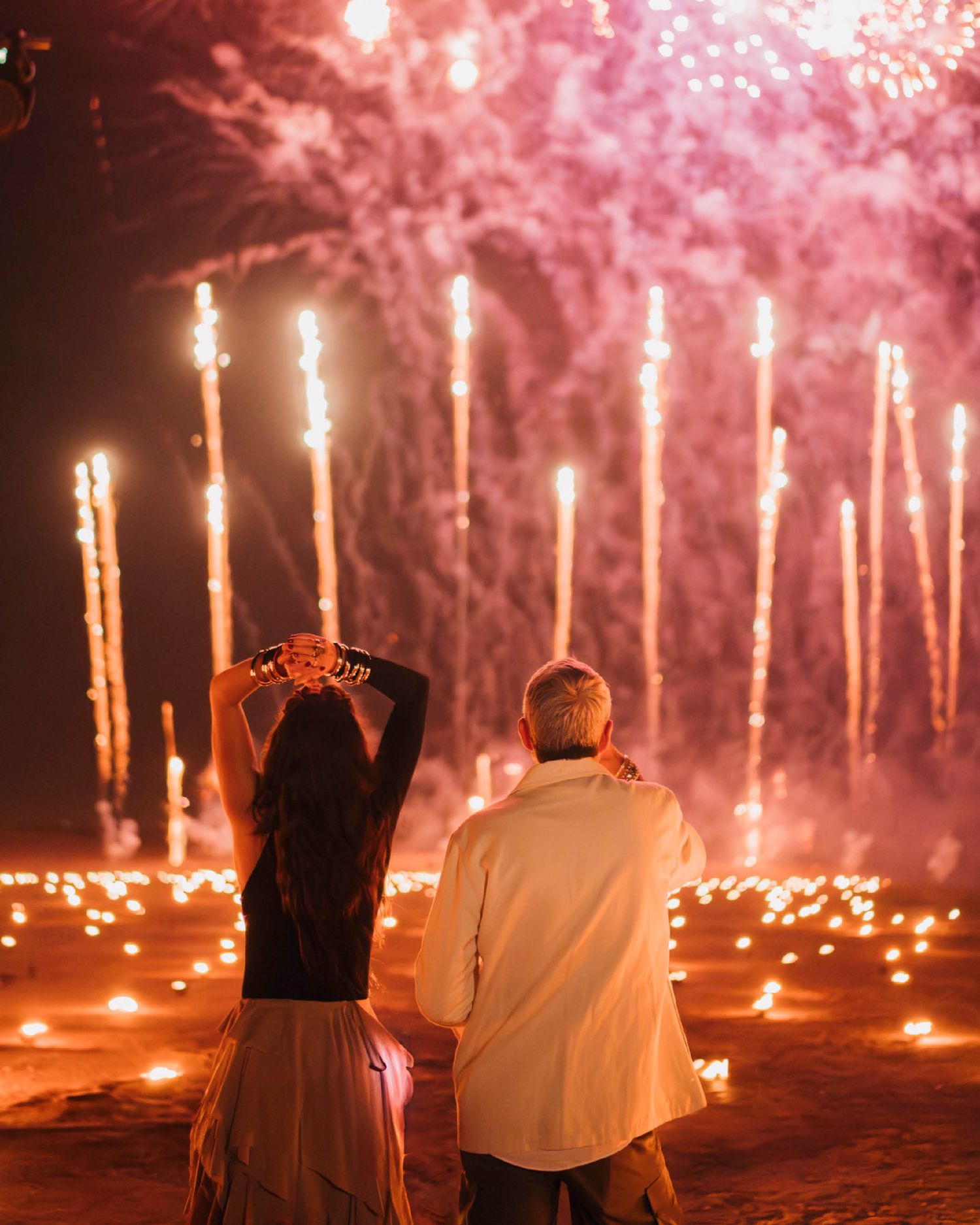 Two people watching spectacular fireworks display with glowing sparklers on the ground creating a romantic atmosphere.