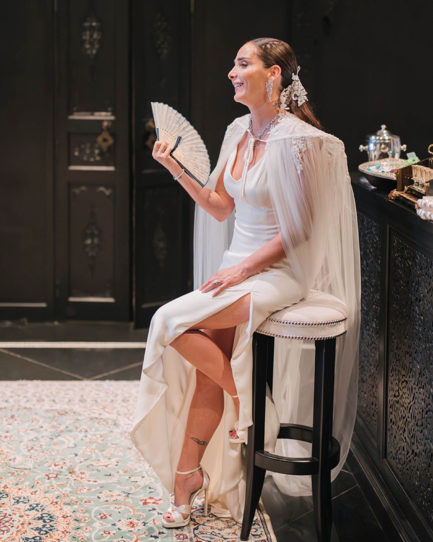 A bride in a white gown sits on a bar stool holding a decorative fan amid confetti-covered floor during a wedding celebration.