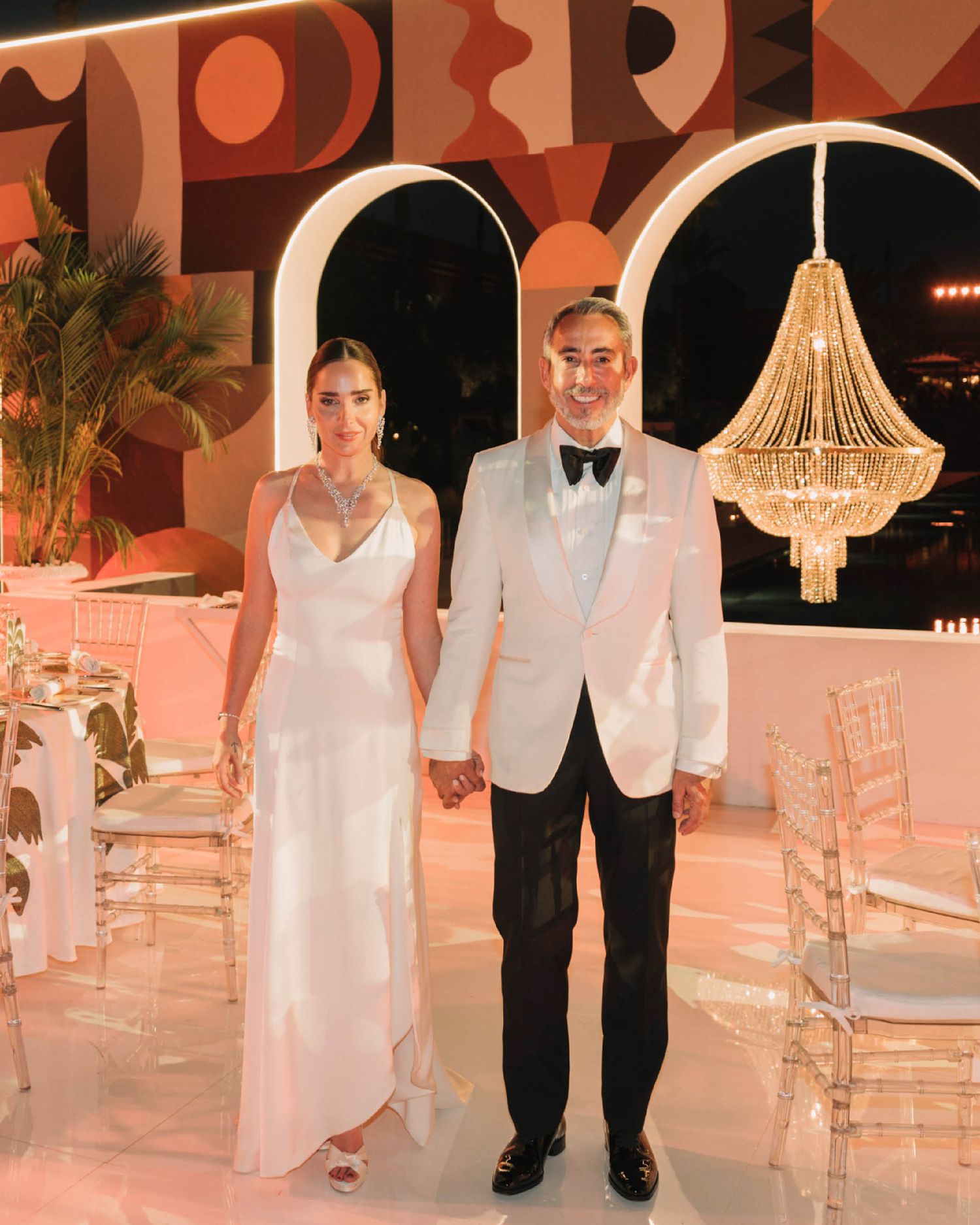 Elegant wedding reception setup with pink decor, white chairs, crystal chandelier and modern art wall in warm tones.