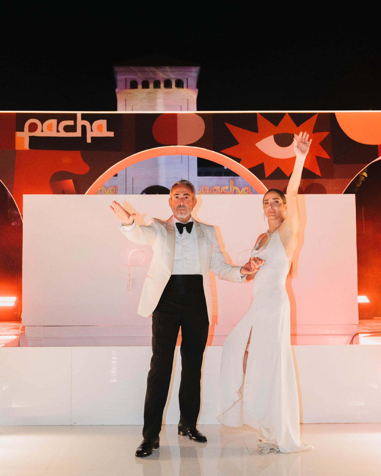 A celebratory wedding moment on a white stage with orange backdrop and decorative lighting.